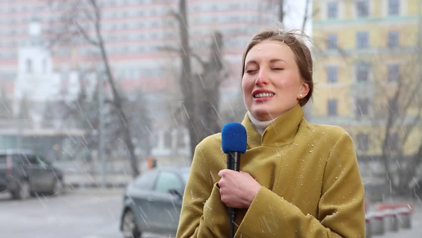 Beautiful blonde girl reporter broadcasts during a snowfall. Concept news, bad weather. The journalist broadcasts on city street.