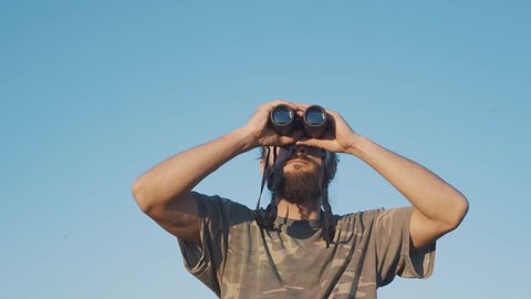 A bearded masculin man in military t-shirt looks through binoculars into the distance. Concept: new horizons, development, travel, territory explorer, adventure time, birdwatching, hunting