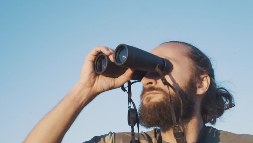 A bearded masculin man in military t-shirt looks through binoculars. Concept: new horizons, development, travel, territory explorer, adventure time, birdwatching, hunting. close up Royalty-Free Stock Footage #1028020952