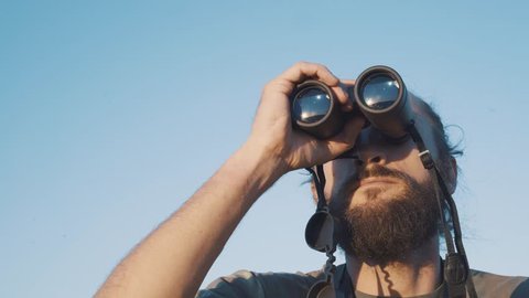 A bearded masculin man in military t-shirt looks through binoculars. Concept: new horizons, development, travel, territory explorer, adventure time, birdwatching, hunting. close up