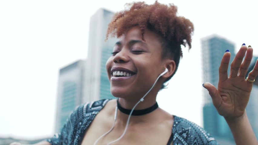 Fashionable Joyful black young woman holding mobile phone listening to music by earphone at Asian city urban background. Slow motion of happy young African woman enjoying music dancing in the street.  | Shutterstock HD Video #1028029667