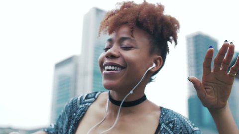 Fashionable Joyful black young woman holding mobile phone listening to music by earphone at Asian city urban background. Slow motion of happy young African woman enjoying music dancing in the street. 