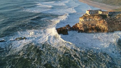 Aerial view of lighthouse on a cliff with a fortress on the coast of the Atlantic ocean with big waves at sunset in Nazare, Portugal, 4k
