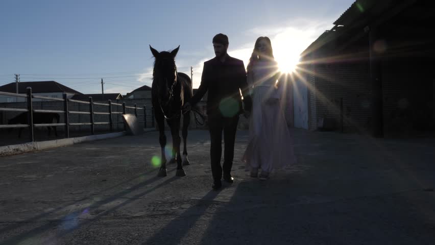 young couple silhouettes with joining hands walk near brown horse against clear blue sky at sunset slow motion