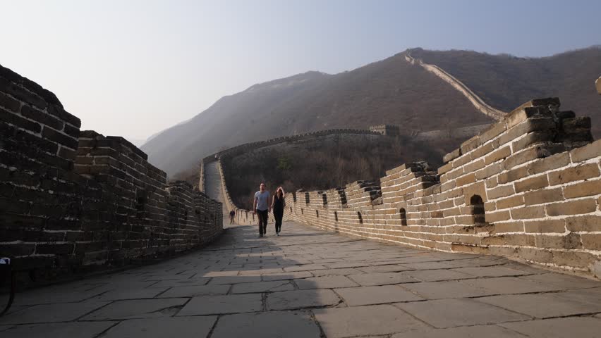 Two tourists walk towards by Great Wall of China, low camera on floor of stone paved wide walkway. People explore famous Chinese landmark, Mutianyu section empty at evening of spring day