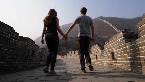 Young man and woman explore Great Wall of China together, low camera on stone pavement of wide passage. Tourists come down holding hands, enjoy empty Mutianyu site at evening time