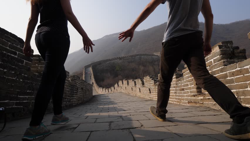 Young woman and man join hands and go along Great Wall of China. Low camera, wide angle shot from floor of stone paved walkway. Tourist pair explore famous Chinese landmark, Mutianyu section