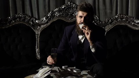 Criminal boss in luxury suit sits on vintage sofa. Crime, mafia, gangster concept. Luxury rich lifestyle.Entrepreneur in elegant suit looks rich. Bearded man champagne glass and smoke cigar