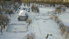 Cinematic drone - aerial footage moving forwards while looking down at dry trees, snowy pathway, cars, a building and people ice skating in a park during winter season.