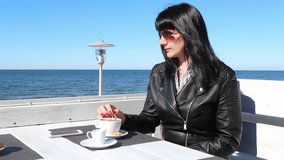 Brunette woman in black leather jacket and sunglasses stirring coffee with a teaspoon in an outdoor cafe near the sea on sunny day