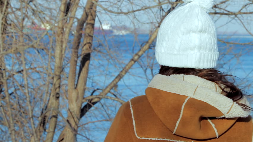 Someone (woman) seen from behind standing by the lake with some dry trees and ships on the background during winter season.