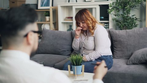 Overweight young woman is crying during conversation with psychologist in office. Upset woman in casual clothing is sitting on couch talking while doctor is listening.