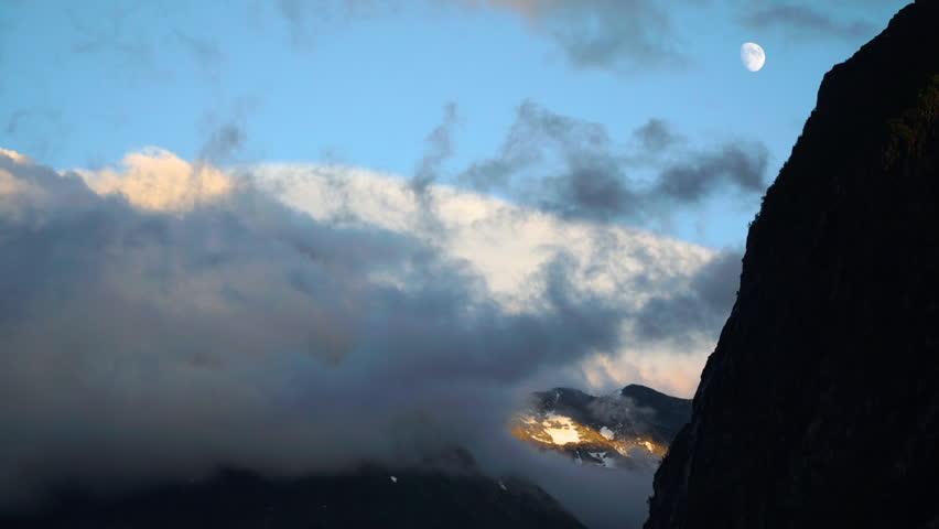 Timelapse of the Norwegian mountains
at sunset with the full moon moving and setting with golden lit clouds floating over a picturesque, ice covered and epic scenery at Jostedalsbreen glacier, Norway.
