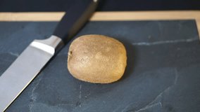 Close up, a woman's hands take a knife and cut the Kiwi exactly in half, then take the two halves and bring it to the video camera, then lay it on the kitchen stone board with a wooden frame