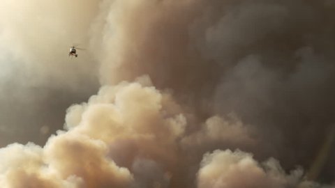 A helicopter flies through a smoky sky as dramatic dark smoke rise up from an out of control wildfire, forest fire, or other disaster.