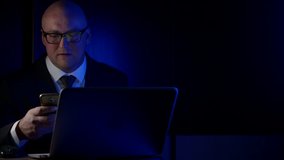 a bald business man, in a suit and glasses, middle-aged, sitting in the dark at a laptop, in the hands of his smartphone.