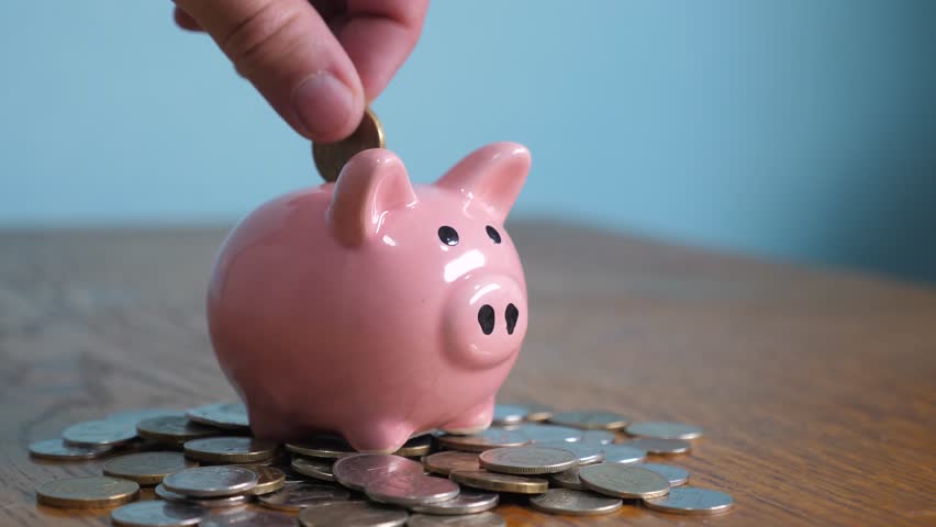 Piggy bank business standing on a pile of coins concept. A hand is putting a coin in a piggy bank on lifestyle a yellow background. saving money is an investment for the future. Banking investment and | Shutterstock HD Video #1028100278