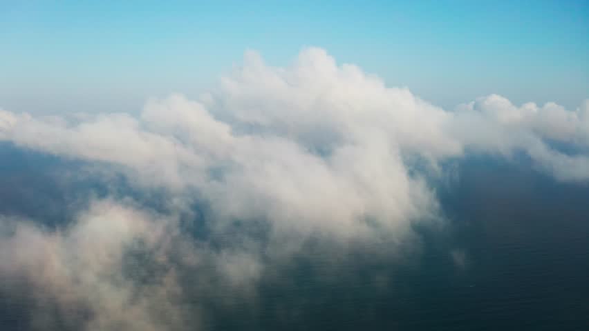 Moving white clouds blue sky on coastline and sea surface scenic aerial view. Drone flies forward high in blue sky through the fluffy clouds on panorama of the sea shore. The sun is hidden. Fog.  Royalty-Free Stock Footage #1028102138