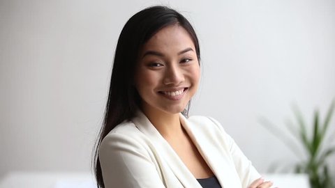 Head shot asian ethnicity millennial businesswoman or company employee standing in office room crossed arms looks at camera laughing feels confident. Team leader, business coach portrait or hr concept