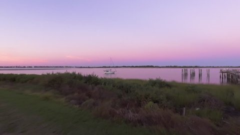 Drone passing slowly over shrubs and a lagoon lake coastline during a serene, evening pastel sky with pilings of an abandoned dock in view, and passing camera-left above a single docked sailboat.