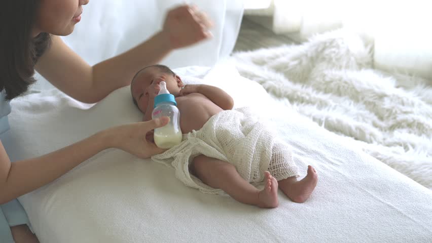 Selective focus Medium shot of young happy mother gently taking care 0-1 month cute newborn baby girl lying down at home, mother feed bottle of milk for newborn, maternity with newborn baby concept