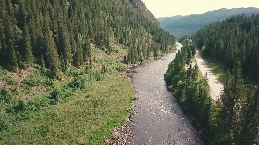 Elevating, high angle forward drone shot over a river next to a road in a forest. Mountains are shown in the bachground. 4k quality video in Norway