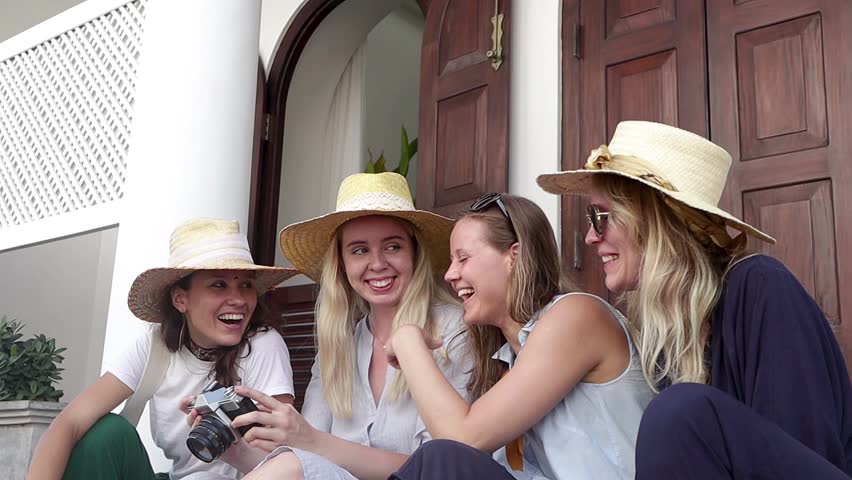 Four young stylish women in straw hats are sitting at the street and watching the photos on the retro camera.