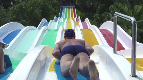 People sliding on colorful water slide in amusement aquapark at summer vacation. People having fun riding on slides in outdoor water park on green palm tree landscape Vídeo Stock