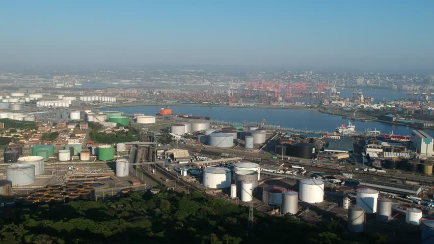 High, wide aerial view of the harbour area of Durban, South Africa. Large chemical and fuel tanks and bright red cranes for loading and unloading ocean going cargo ships in the distance. Drone view.