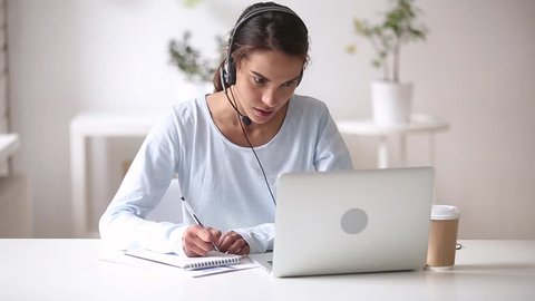 Woman sitting at desk wearing headset talking looks at laptop screen noting in notepad learn foreign language using online course, call center employee give support distantly to company client concept