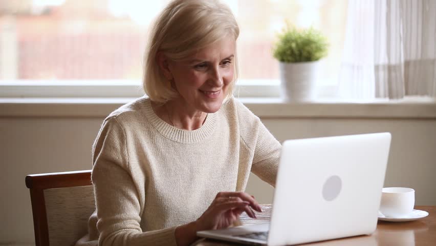 Smiling elderly blond woman sitting at table using computer communicating online with grown up children, chatting with grandchildren or friends via internet at home surfing website, new apps concept Royalty-Free Stock Footage #1028137064
