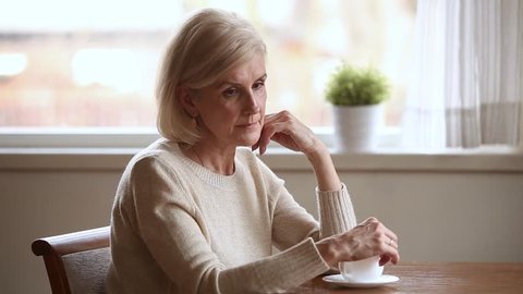 Lonely disappointed elderly blond woman sitting at table with coffee cup lost in sad thoughts feels upset, mature alone melancholic grandma reminiscing relive the past memories regrets in life concept