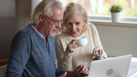 Happy elderly blond wife drinks coffee chatting with grey haired husband mature family sitting at table in kitchen use laptop discuss new app browse website, buying online, internet shopping concept