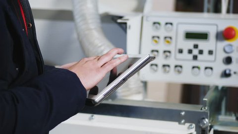 Factory worker are setting up single sided automatic edge bander machine on the factory. A young man is holding a digital tablet, calibrating a modern machine. Close-up video