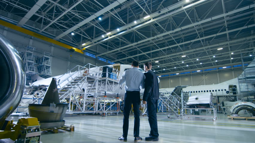 Aircraft Maintenance Mechanic and Chief Engineer Have Discussion, Consult Blueprints While Standing in a Big Airplane Development Facility. They Analyze, Inspect, Develop and Design Airplanes