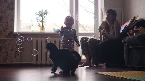 a young beautiful woman blowing soap bubbles inside the room with big window on background and a little baby boy and a black cat are catching this bubbles in slow motion 4K video