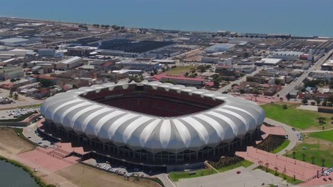 Port Elizabeth, South Africa - circa 2010s: Aerial fly forward, tilt down onto Nelson Mandela Bay Stadium near ocean and lake. See unmaintained green soccer pitch between red seats and open white roof