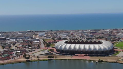 Port Elizabeth, South Africa - circa 2010s: Nelson Mandela Bay Stadium. Wide aerial view, turning around stadium that lies between a lake and the ocean. Large container ships out at sea