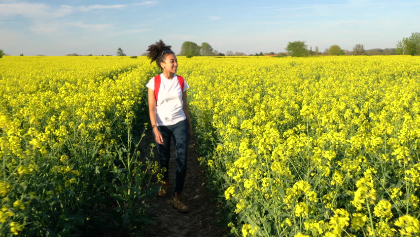 Beautiful happy mixed race African American girl teenager female young woman hiking with red backpack on path through field of yellow flowers