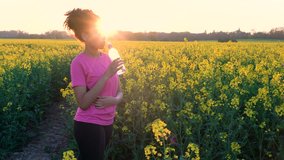 4K video clip of beautiful healthy mixed race African American girl teenager female young woman running or jogging and drinking a bottle of water in field of yellow flowers