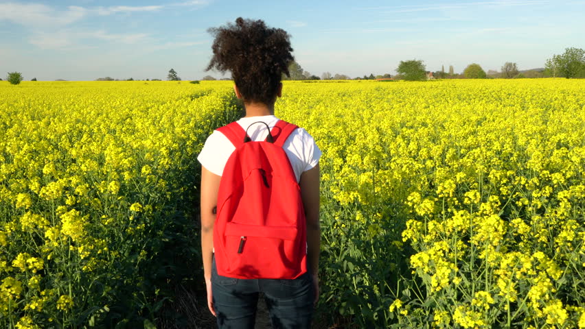 Beautiful happy mixed race African American girl teenager female young woman hiking with red backpack on path through field of yellow flowers