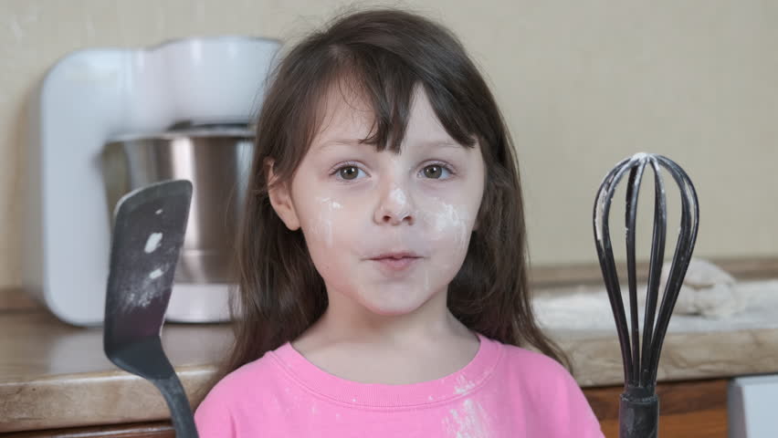 Funny child in the kitchen. A girl soiled in flour shows her tongue.