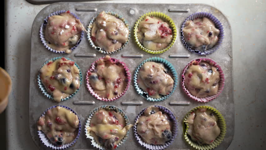 Pouring sugar over muffin batter with tasty fruit and colorful wrappers in metal baking tray overhead