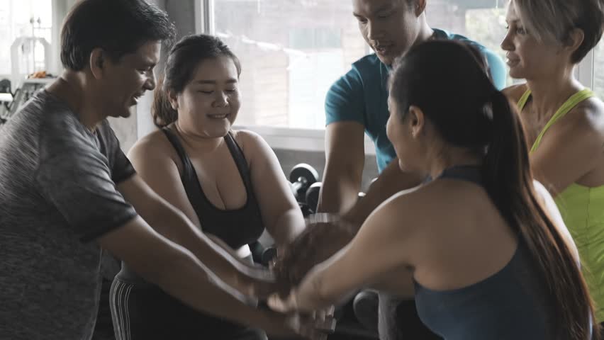 Group of young people in gym putting hands together. Team work and accomplishment concept, mix races of asian and caucasian man and woman, young and senior.