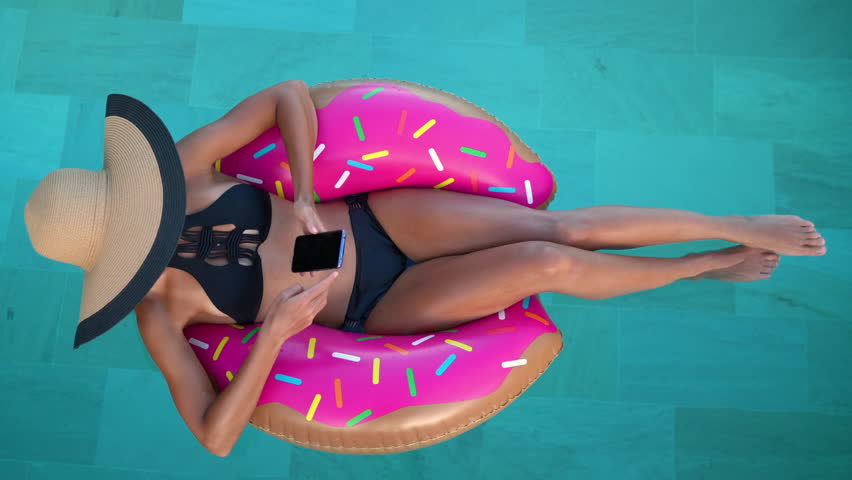 Summer Vacation Woman in bikini on inflatable donut mattress using mobile cell phone in swimming pool. Top view of girl relaxing sunbathing enjoying travel holidays at resort pool. Luxury lifestyle.