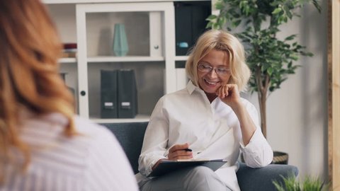 Positive psychoanalyst blond woman is smiling and laughing during conversation with female patient in office room. People, dialogue and psychology concept.