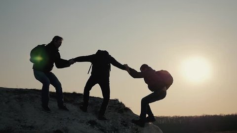 Silhouettes of tourists climbing mountain cliffs against the backdrop of a sunset, helping each other's hand. Help in the mountains and teamwork in hiking. Teamwork concept.