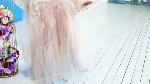 A young girl wearing tulle skirt sits in a hanging lounger at a photo shooting and dangles her feet in high heels.