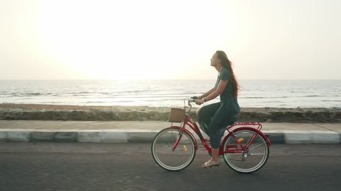 Smiling red hair female in green dress ride vintage bicycle along coast line 4k