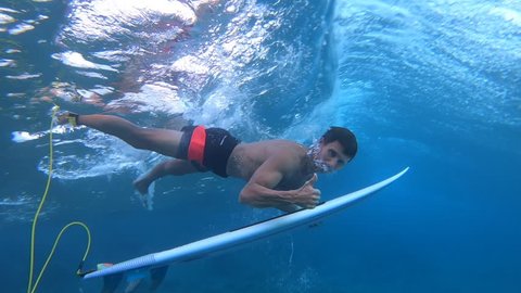 Surfer dives under the wave with the surfboard (the trick named in surfing as Duck Dive) looks into the camera and shows the thumb up